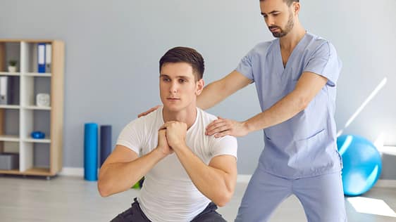 Physiotherapist Helping Young Athlete Do Squats during Rehabilitation Therapy after Sports Injury