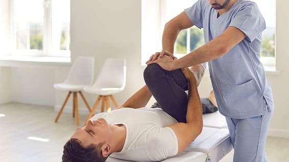 Serious Doctor Helping Male Athlete Do Physical Exercise during Rehabilitation after Leg Injury